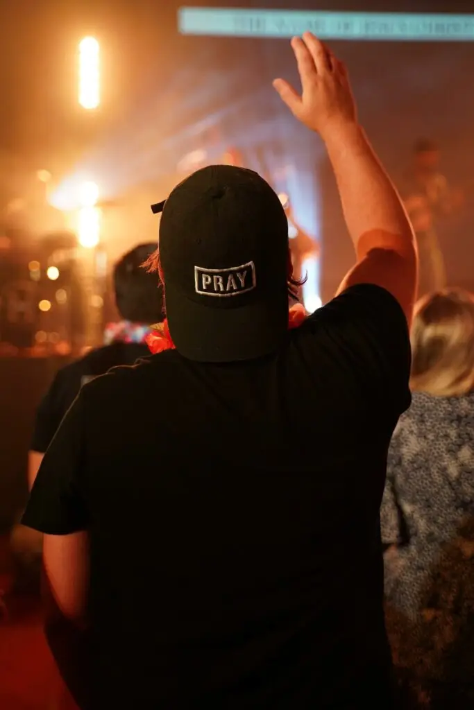 An image of the back of a man raising his hand in worship at church, wearing a backwards hat that reads 'pray', representing Dare 2 Share's blog titled 6 Essentials for Youth Leader Training.