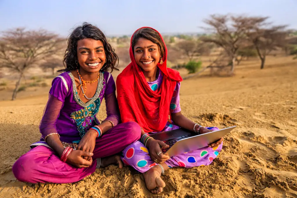 Happy Indian young girls sitting on a sand dune and using laptop in desert village, Thar Desert, Rajasthan, India.