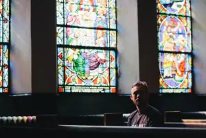 Picture of a man sitting in a church pew with stained glass behind him